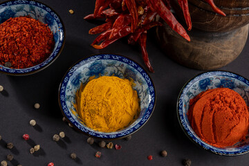 Close up on three blue bowls with whole, granular and powdered red pepper and yellow mixed ginger, curry, turmeric powder and peppercorns. Oriental food seasoning herbs on black background