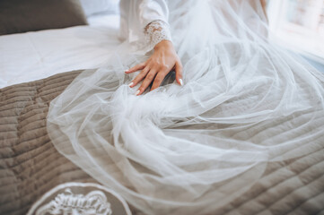 The bride in a white coat, lingerie sits on a sofa with a long veil and gently holds her hand....