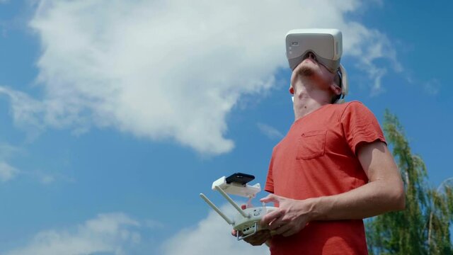 Man controls a quadrocopter through a remote control and looks at the video with goggles on his head. Drone operator in red T-shirt in a park shoots video on drone. Blue sky on the background