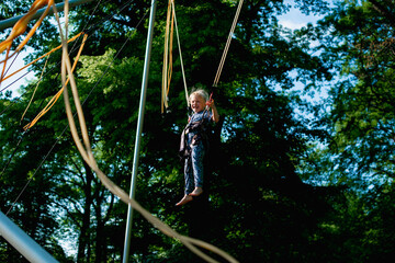 The girl is jumping on a bungee trampoline. A child with insurance and stretchable rubber bands hangs against the sky. The concept of happy childhood and games in the amusement park