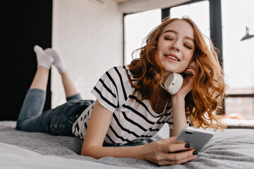 Attractive female model with phone in hand lying on bed. Happy relaxed girl enjoying music in headphones.