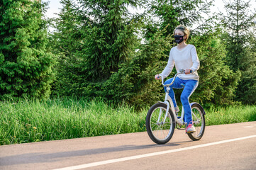 Teenager girl in a protective mask rides a bicycle in a city park. Copy space
