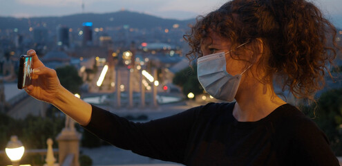 Tourist woman with mask on her face takes a selfie at night in the city. New normal	