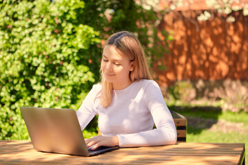 A cute young teen girl is learning online on a laptop while sitting at a wooden table in the back garden of a house.