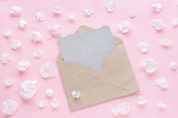 Craft envelope with empty card on pastel pink background and white flower arrangement. Flat lay, top view.
