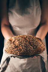 Female hands holding loaf of homemade rye bread with sesame and linseeds on towel. Concept for bakery poster or cover