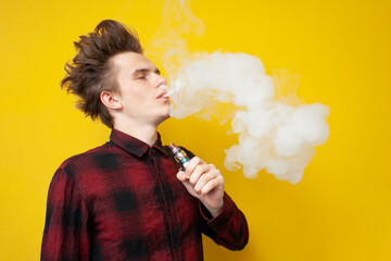 hipster guy smokes vape on yellow isolated background, man exhales a cloud of smoke and holds an...