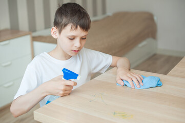 A school-aged boy cleans the room, puts detergent on the table. In his hand he holds a blue cloth...