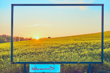 A sunset over a rapeseed field, viewed through a frame. Under the frame is an arrow pointing to the right, indicating a distance of 500 meters.  