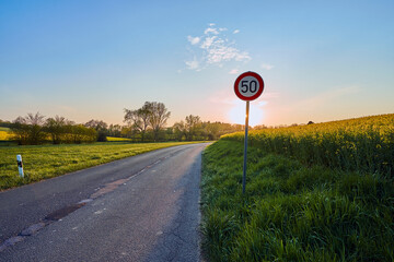 A country road with rape fields and meadows along the road. A red and white traffic sign with the...