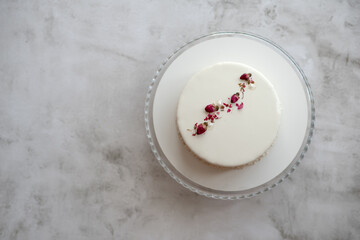 Obraz na płótnie Canvas Small mousse cake with smooth white glaze decorated with dried rose buds in concise simple design on gray textured background. Place for text