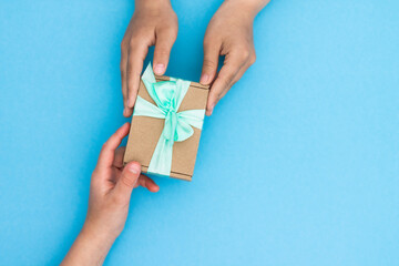 Hands giving or receiving a gift in craft paper with a mint ribbon on blue background. Top view. Copy space.