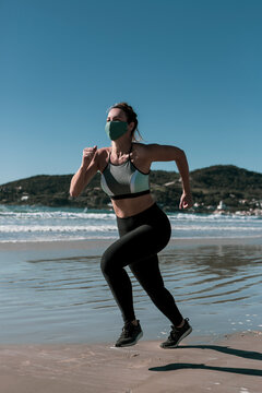 Beautiful athletic young woman running on the beach with green hills on the background wearing mask and sportswear on a sunny day and blue sky during covid-19 pandemic