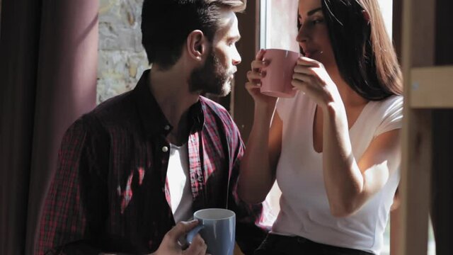 Loving couple drinks morning coffee while standing by window preparing to work and discussing plans. Wife is sitting on windowsill with a coffee while a loving husband is standing nearby. Prores 422.