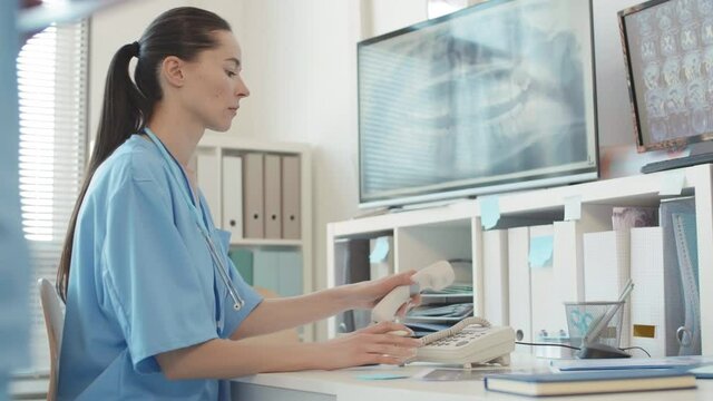 Medium shot of young Caucasian female doctor wearing uniform sitting at desk in office, writing in patient card and then answering phone call