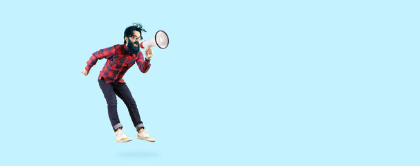 Bearded hipster man in glasses, jumping and shouting in megaphone, photo on blue background
