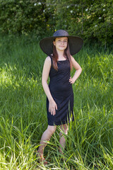 Attractive young girl in a black dress and hat in a summer green park.