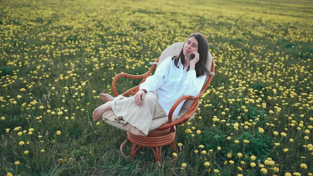 Beautiful attractive woman sitting in a chair on the field of dandelions. She is relaxing and speak by phone. Happy woman is laughing. Enjoy the moment.