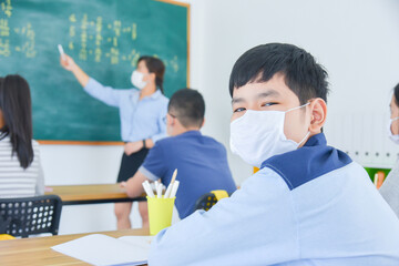 Young asian schoolboy wearing facial mask sitting in classroom with classmate looking at camera while teacher teaching on background.