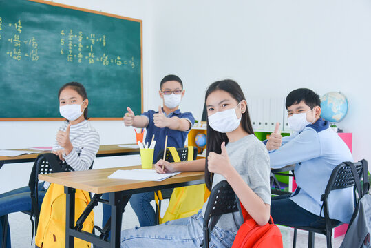 Group of young asian students wearing facial mask sitting in classroom with classmate ,everyone looking at camera and showing thumb up. - Powered by Adobe