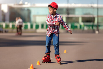 Cheerful little african american boy on rollers skates in summer park at asphalt road between...