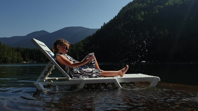 Kid girl lie on a sunbed in sunglasses and a boho silk shawl. Child rest on a flood wood underwater pier. The pavement is covered with water in the lake. In the background are mountain and a forest.
