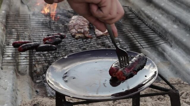 Una mano de un chico saca chorizos cocinados de la parrilla a un plato.