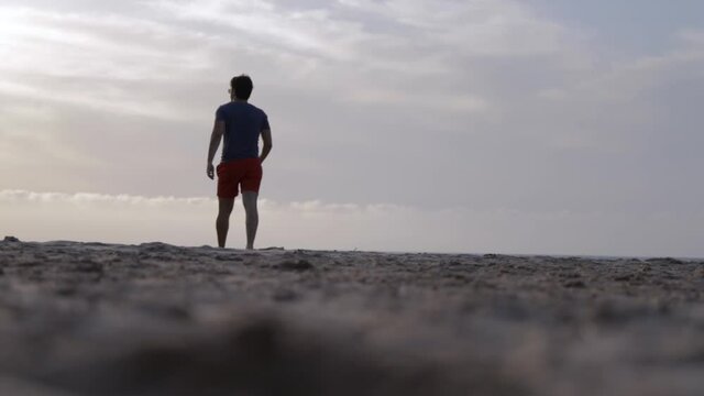 Low Angle Panning Shot Of Man Walking Across Sandy Beach Away From Camera