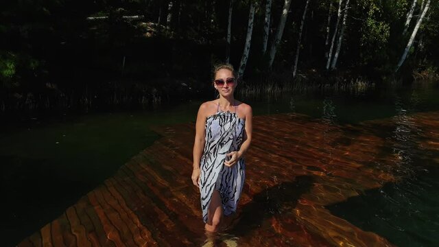 Woman walk on water on pier in sunglasses and a boho silk shawl. Girl rest on a flood wood underwater dock. The pavement is covered with water in the lake. In the background are mountain and a forest.