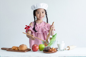 Cute Asian girl with chef hat and salad bowl for breakfast, holding wooden spoon and smiling looking at camera