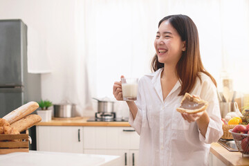 Obraz na płótnie Canvas Young Asian woman in the kitchen drinking milk for breakfast and bread in her hand, Healthy food concept