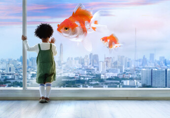 Dark skinned boy looking out of window, gold fish flying through the air over the city with...
