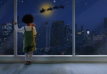 Fototapeta na wymiar Dark skinned boy looking out of window, Santa Claus riding on sleigh against bright sky over city, Merry Christmas and Happy New Year concept