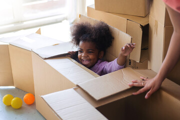 Dark skinned child girl playing in cardboard box in room, hiding in a box while playing hide and...