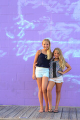 A portrait of two sisters with long blonde hair with purple and white backdrop.  