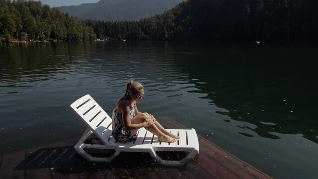 Woman lie on a sunbed in sunglasses and a boho silk shawl. Girl rest on a flood wood underwater pier. The pavement is covered with water in the lake. In the background are mountain and a forest.