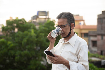 Portrait of an Indian old man with corona preventive mask watching his cell phone while drinking coffee on rooftop during afternoon in home isolation.Indian lifestyle, disease and home quarantine.