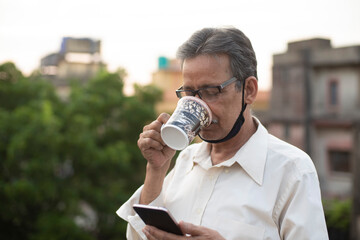 Portrait of an Indian old man with corona preventive mask watching his cell phone while drinking coffee on rooftop during afternoon in home isolation.Indian lifestyle, disease and home quarantine.