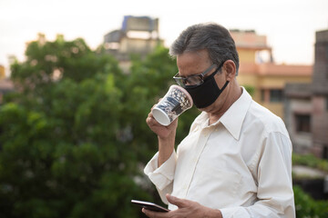 Portrait of an Indian old man with corona preventive mask watching his cell phone while drinking coffee on rooftop during afternoon in home isolation.Indian lifestyle, disease and home quarantine.