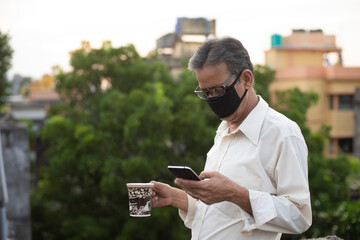 Portrait of an Indian old man with corona preventive mask watching his cell phone while drinking coffee on rooftop during afternoon in home isolation.Indian lifestyle, disease and home quarantine.