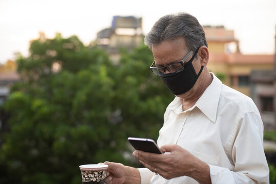 Portrait of an Indian old man with corona preventive mask watching his cell phone while drinking coffee on rooftop during afternoon in home isolation.Indian lifestyle, disease and home quarantine. - Powered by Adobe