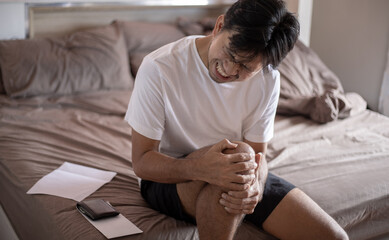 young asian man holding knee in bed room, man hurt pain at knee concept at home.