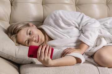 A smiling girl in home white coat, with phone in her hands, lies on sofa.