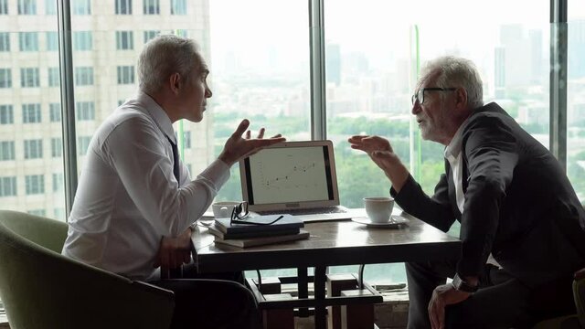 two senior management businessman debating  in meeting with data information in laptop. old man ceo discussing with client in cafe. serious mature manager talking by window over cityscape hi class