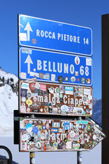 Passo Fedaia, Italy, 02/21/2020 , Sign on Passo Fedaia indicating directions to Rocca Pietore, Belluno, and Malga ciapela, completely covered with stickers left by tourists as memorial.