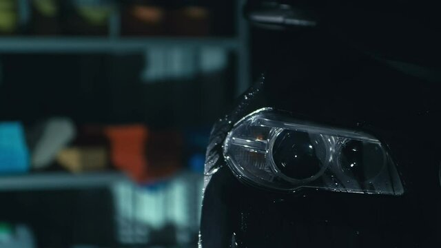 Car headlight wash. Washing modern vehicle body by high pressure jet wash hose water. Auto glass headlamp, angel eyes in drops. Close up of the spotlight of a black automobile as it gets cleaned.