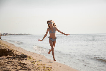 Fototapeta na wymiar Happy preteen girl running on the sandy beach. The kid dressed swimsuit and sunglasses