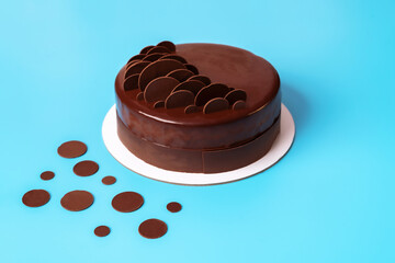 Dark chocolate mousse cake covered with mirror glaze and decorated with round chocolate chips on blue background. Modern stylish cake. Close-up. Copy space.