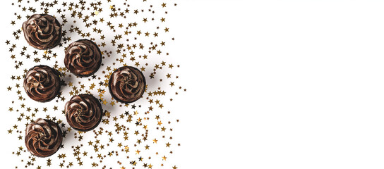 Three delicious chocolate cupcakes with chocolate icing and sprinkled gold sparkles on white background with golden star shaped confetti. Holiday concept. Web banner. Flat lay style. Place for text.