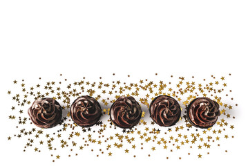 Chocolate cupcakes with chocolate icing and sprinkled gold sparkles on white background with golden star shaped confetti. Holiday concept. Backdrop for your design. Flat lay style. Place for text.
